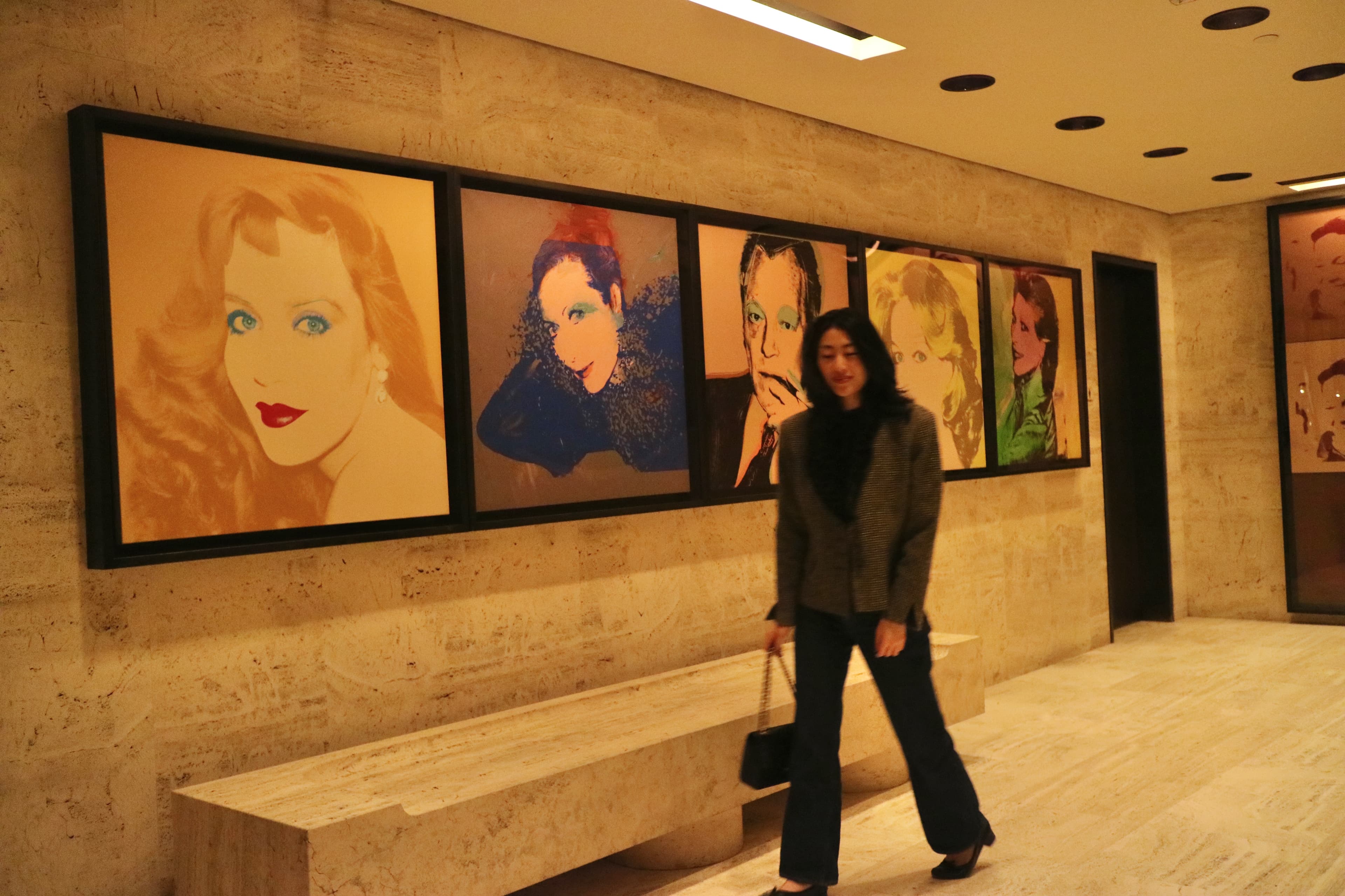 Isabelle Yang walking past a row of framed Warhol-style portraits in a warm-lit interior